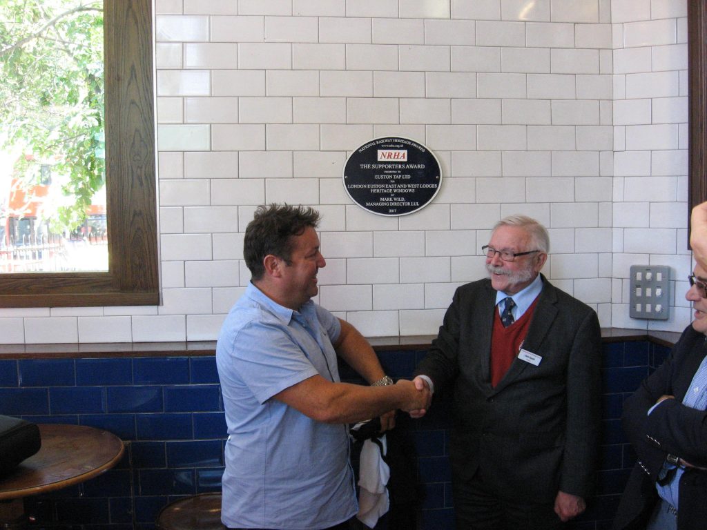 Attendees at meetings often long for the end of the debate but committee members of the National Railway Heritage Awards had a special reason for anticipating the end of the trust’s committee meeting on Wednesday 26 September as the date had been set for the official unveiling of the plaque made to Euston Tap Ltd for the restored windows in the two lodges at Euston. The plaque, presented at the annual ceremony held at Merchant Taylors’ Hall on 6 December 2017, recognised the project to restore the windows of the late 19th century lodges being given the Supporters Award. As was said at the ceremony ‘It has been a thoroughly worthwhile enterprise for which all involved can take great credit.’ 	One of the more entertaining facets of the event was the ‘curtain’ used to hide the plaque before its unveiling by Chris Smyth, from the Heritage Railway Association (one of the award’s sponsors) – this was one of the T-shirts normally worn by the staff behind the bar.